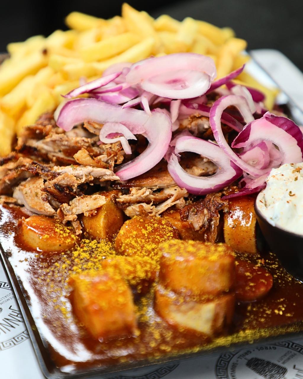 Ein Teller mit Pommes, Gyrosfleisch, Zwiebeln, Currywurst und cremigem Dip