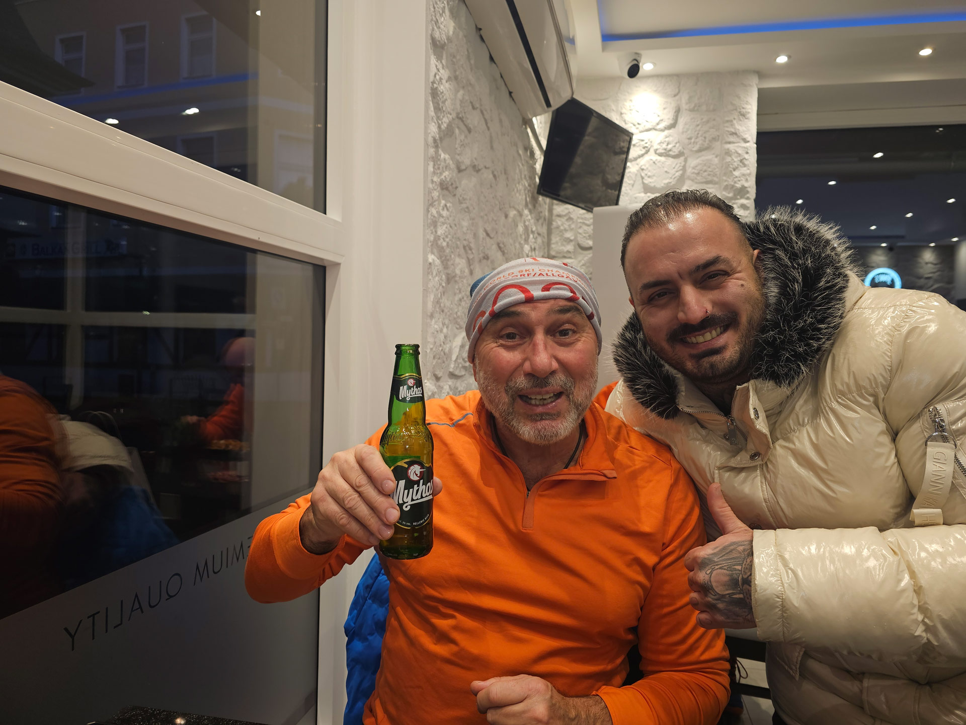 Zwei lachende Männer in einer Bar, einer hält eine grüne Flasche und trägt Orange