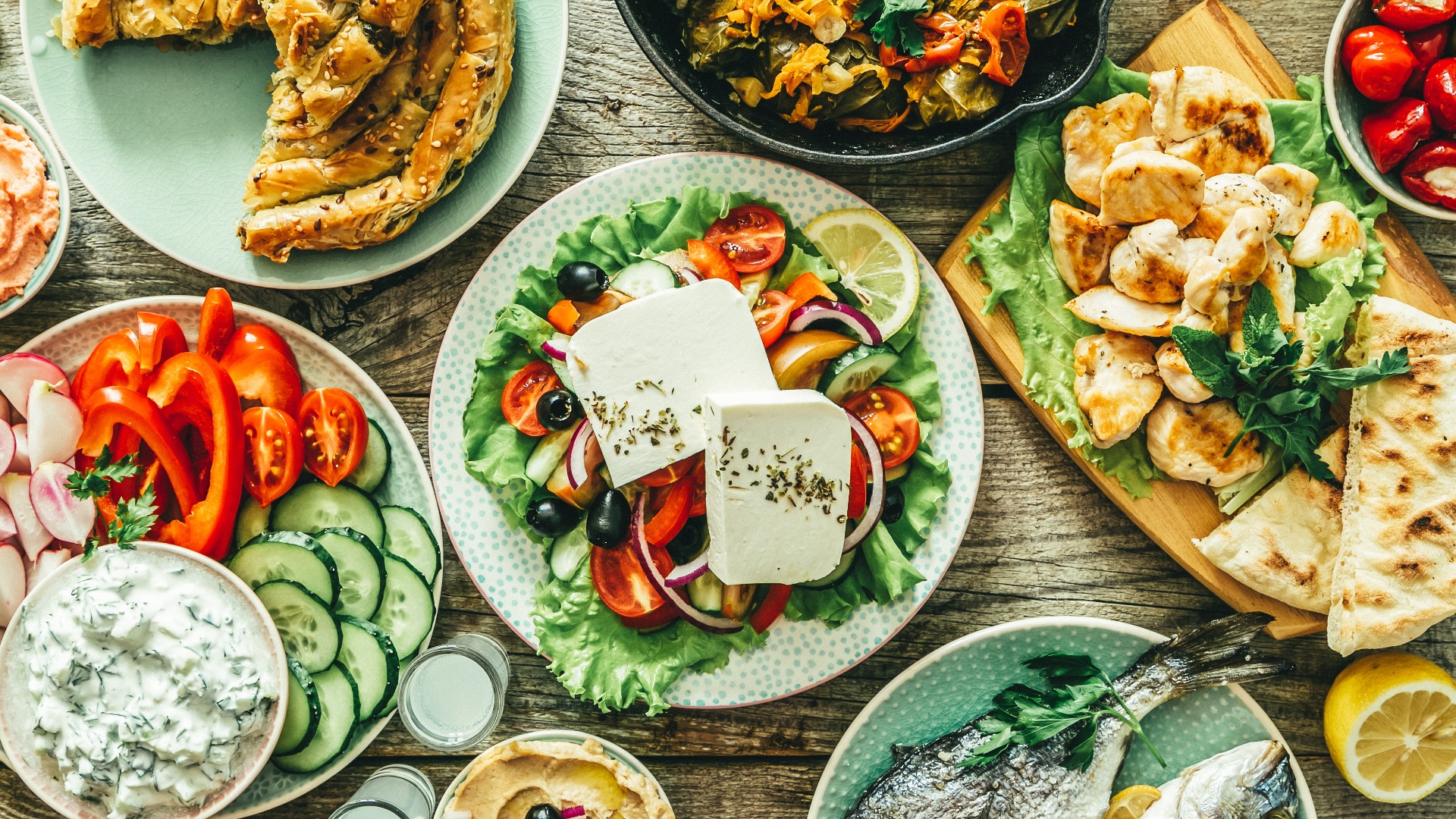 Auswahl an traditionellen griechischen Speisen – Salat, Meze, Pastete, Fisch, Tzatziki, Dolma vor einem Holzhintergrund