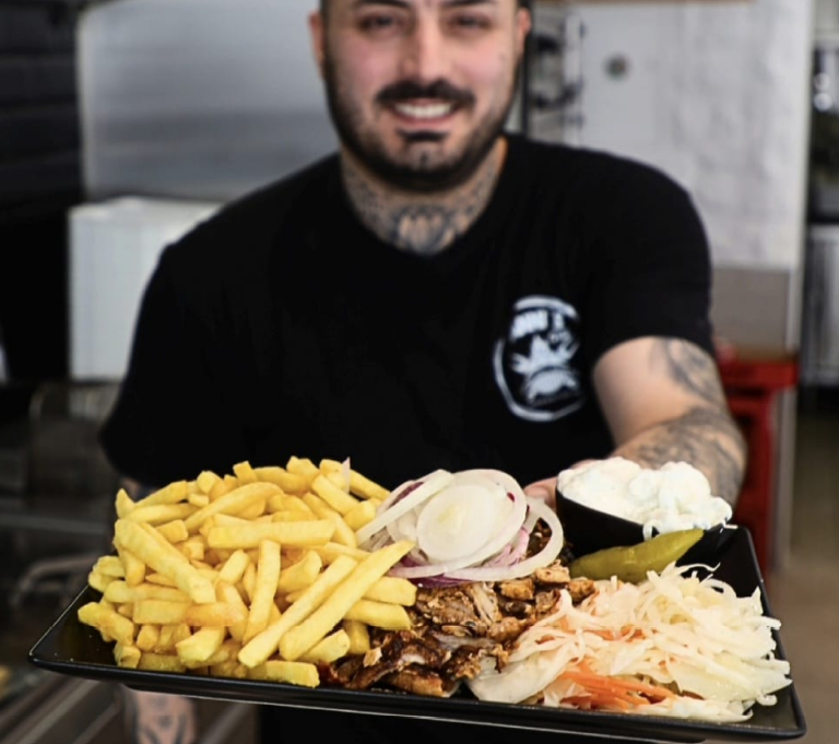 Ein tätowierter Mann in schwarzem Shirt hält einen Teller mit Pommes und Fleisch